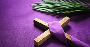 A wooden cross with a purple drape is flat on a cloth of purple with a palm frond in the upper right corner.